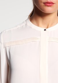 Blusa rosa chiaro con una texture liscia, caratterizzata da pieghe verticali sul petto e un collo rotondo, chiusa con un piccolo bottone.