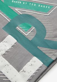 Tessuto grigio con motivi geometrici in verde e bianco, con dettagli di cucitura, testo con marchio e una finitura texture.