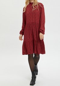Röd mönstrad klänning med långa ärmar och ett lager på lager kjol, med en volangsågad halsringning och subtila svarta triangelmönster. Svarta ankelskoboots.