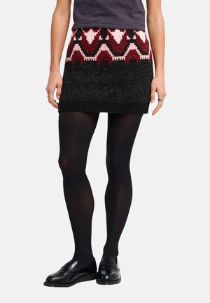 Personne portant une mini-jupe en maille noire à motifs avec des détails rouges et roses, des collants noirs et des mocassins noirs, les mains le long du corps.