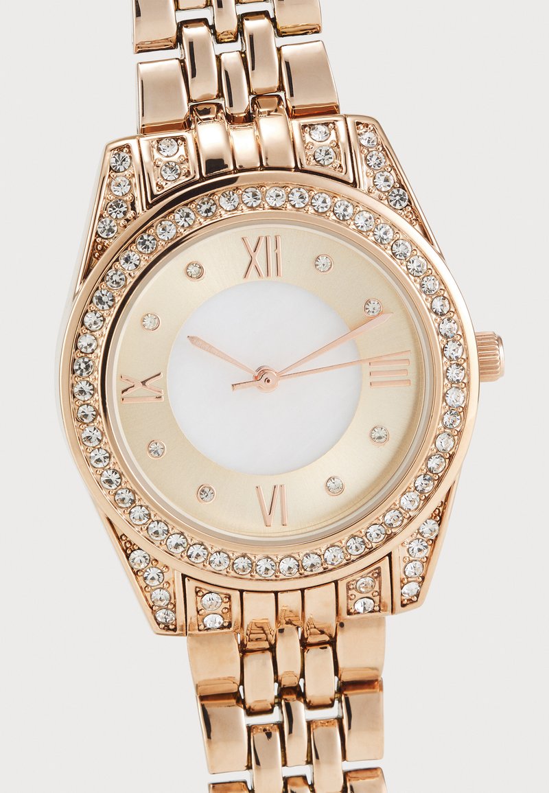 Montre dorée avec un cadran rond, un centre blanc, des chiffres romains et des accents en strass clairs sur la lunette. Design de bracelet en maillons métalliques.