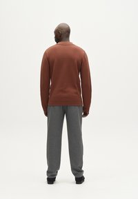 Maglione marrone a maniche lunghe con una texture sottile, abbinato a pantaloni grigi larghi. Lo sfondo è bianco semplice.