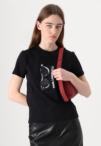 Juodas medvilnės marškinėliai su blizgiais baltais saulės akinių grafikos ir "KARL LAGERFELD" tekstu; derinami su raudona rankine.