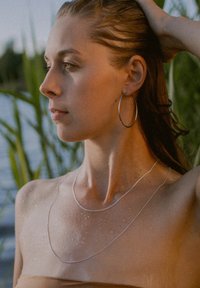 Silver hoop earrings and layered silver necklaces adorn a woman with wet, glistening skin, standing against green foliage by water.