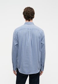 Lichtblauw overhemd met lange mouwen, voorzien van verticale witte strepen. Gemaakt van geweven stof met een standaard kraag en knoopmanchet.