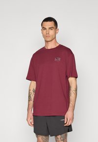 Bordeaux bomulds T-shirt med rund hals og korte ærmer, prydet med et farverigt grafisk tryk på venstre brystområde. Casual pasform.