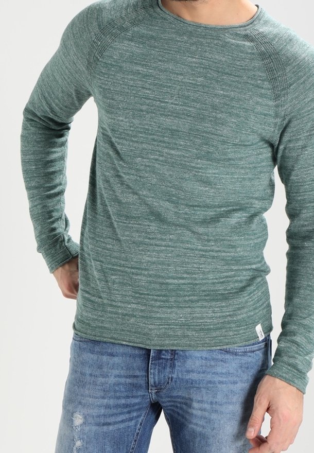 Homme portant un pull vert à manches longues et un jean bleu délavé, debout avec une main dans la poche devant un fond uni.