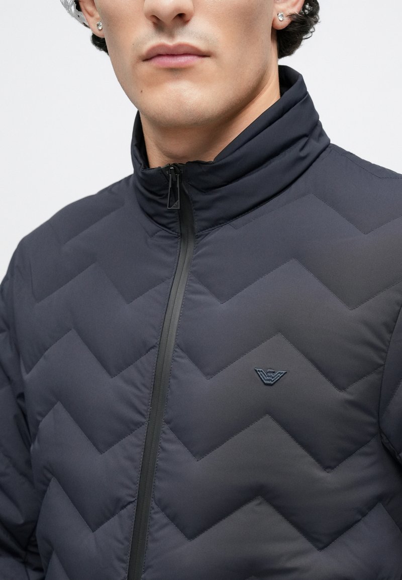 Doudoune bleu marine avec un motif matelassé en chevrons, col montant et fermeture éclair. Présente un logo discret sur la poitrine.