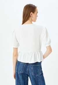 Blusa bianca texturizzata con maniche a sbuffo corte e vita a peplo, abbinata a jeans in denim blu con un motivo sottile e tasche posteriori.
