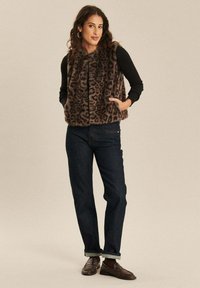 Vrouw in een leeuwenprint neppelzen vest over een zwart langemouwen shirt, donkere jeans, bruine loafers, staand met handen in de zakken.