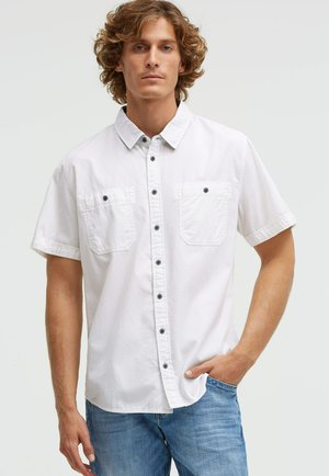 Young man with curly hair wearing a white short-sleeve button-up shirt and blue jeans, one hand in pocket, neutral background.