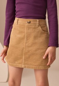 Mini gonna in velluto a coste color beige con righe verticali, dotata di chiusura con bottoni frontale e due tasche laterali. La modella indossa una maglietta a maniche lunghe viola.