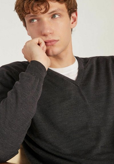 Pull en V gris foncé en tissu texturé, avec des manches longues et une coupe décontractée, superposé à une chemise blanche. Détails de design simples.