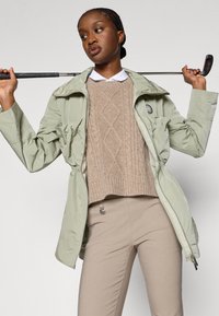 Φωτεινό πράσινο αντιανεμικό με λαμπερή υφή, με ζωνάκι στη μέση, φο�ρεμένο πάνω από πλεκτό πουλόβερ σε μπεζ απόχρωση και μπεζ παντελόνι. Κρατά χαλαρά μία μπαστούνι του γκολφ.