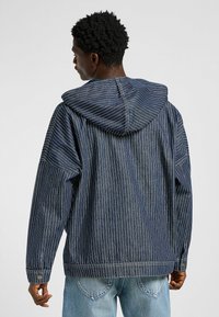 Jeansjacke mit Kapuze, die ein blau-weißes vertikales Streifenmuster aufweist, lange Ärmel und eine lockere Passform hat. Detaillierte Nähte an den Bündchen.