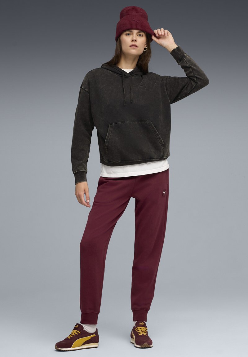 Svart, oversized hoodie med en ficka framtill, vit t-shirt under, vinröda sweatpants och en vinröd stickad mössa. Mörka sneakers med gula detaljer.