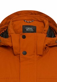 Pomarańczowa kurtka puchowa z kołnierzem i czarnymi elementami metalowymi, z metką z logo i detalami w szyciu. Materiał wydaje się być teksturowany i wytrzymały.