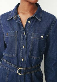 Robe chemise en denim foncé avec un col, des accents de boutons dorés, des poches poitrine et une ceinture assortie avec une boucle circulaire.