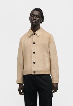 Jeune homme à la peau foncée et aux dreadlocks courtes, portant une veste boutonnée en daim beige et un pantalon noir sur un fond blanc uni.
