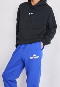 Černá mikina s kapucí a světle modrým logem Nike, spárovaná s modrými tepláky, které mají bílé nápisy "nike sportswear" a elastický pás.
