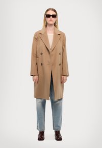 Beige double-breasted coat with large buttons, worn over a light top and blue jeans; paired with dark sunglasses and brown boots.