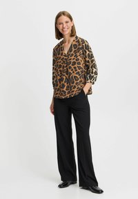 Blouse en imprimé léopard marron et noir, décolleté en V, coupe ample, manches trois-quarts ; portée avec un pantalon large noir et des chaussures noires.