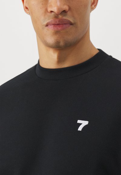 Sudadera negra con cuello redondo y puños acanalados, que presenta un pequeño logo blanco bordado con el '7' en el pecho. Textura de tela suave.