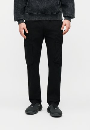 Mann trägt schwarze Cargohosen und schwarze Slip-On-Schuhe, steht vor einem schlichten weißen Hintergrund.