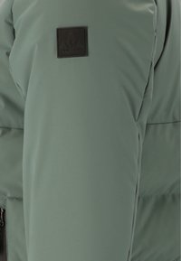 Giacca puffer verde con superficie testurizzata, chiusura con zip, design imbottito e patch con logo nero sul braccio superiore.