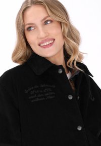 Smilende blond kvinde med bølget hår iført en sort fløjlskjortjakke med trykknapper og broderet tekst på brystet, kigger til venstre.
