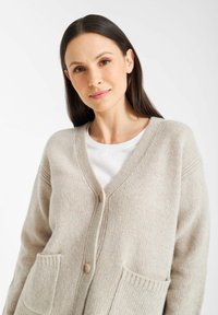 Beżowy sweter zapinany na guzik z dekoltem w serek, w oversize'owym fasonie, z dwiema przednimi kieszeniami. Miękka faktura z prążkowanymi akcentami.
