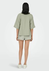 Helles grünes, locker sitzendes Oberteil mit Falten auf der Vorder- und Rückseite, kurzen Ärmeln, kombiniert mit passenden Shorts. Das Model trägt weiße Sneaker.