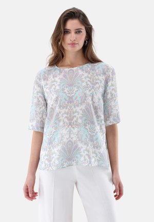 Frau mit langen braunen Haaren, die eine pastellblaue und lavendelfarbene Bluse mit Blumenmuster sowie weiße Hosen trägt, steht vor einem weißen Hintergrund.