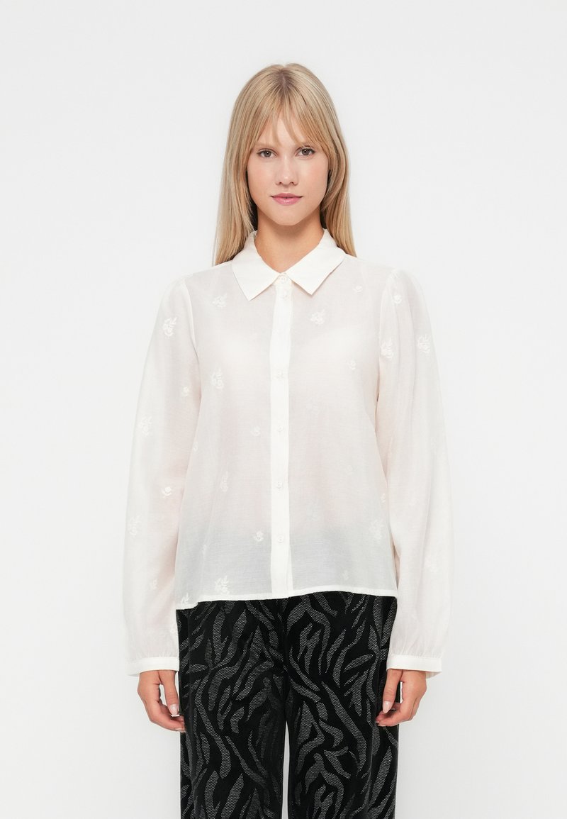 Blusa bianca con bottoni, maniche lunghe, colletto e dettagli floreali ricamati su un tessuto trasparente, abbinata a pantaloni neri con motivo.
