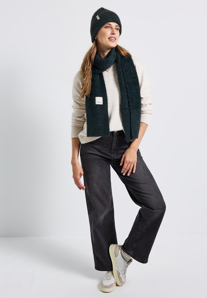 Tmavě zelená pletená čepice a šátek, krémový svetr, černé kalhoty s širokými nohavicemi a světlé tenisky s texturovaným povrchem a drobnými logy.