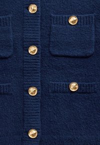 Tamsiai mėlynas megztinis su tekstūruota medžiaga, puoštas auksiniais apvaliais mygtukais ir dviem priekinėmis kišenėmis su ribsčių detalėmis.