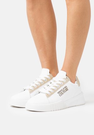 Baskets blanches basses avec des détails en paillettes dorées et le logo "Versace Jeans Couture" portées aux pieds sur un fond uni.
