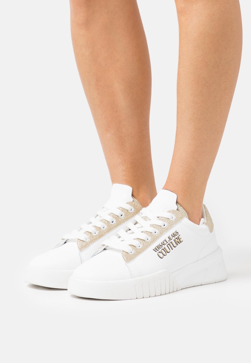 Baskets blanches basses avec des détails en paillettes dorées et le logo "Versace Jeans Couture" portées aux pieds sur un fond uni.