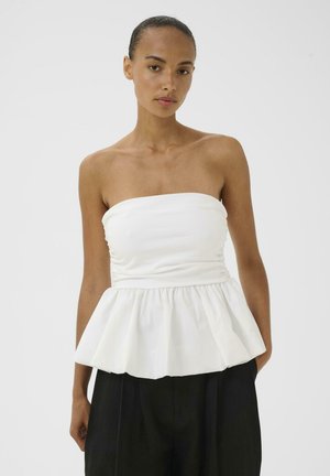 Kobieta w białym, bezramkowym topie typu peplum, zestawionym z wysokim stanem czarnych spodni, na prostym tle.