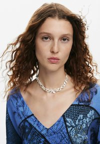 Collana choker in argento con accentuazione floreale indossata sopra un maglione a piccole fantasie blu con scollatura a V profonda. Presenta ricci naturali e leggeri lentiggini.