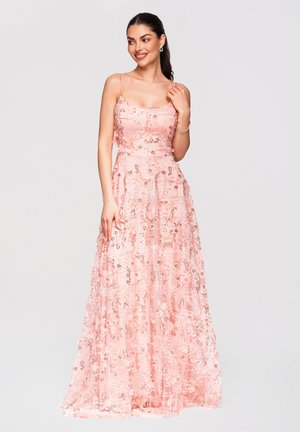 Femme en robe rose sans manches, longue jusqu'au sol, avec des broderies florales, souriante et posant avec une main près de son cou.