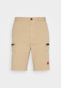 Pantalones cortos de carga beige hechos de un tejido resistente, con dos bolsillos laterales con cremalleras, un cierre de botón y una etiqueta de acento roja.