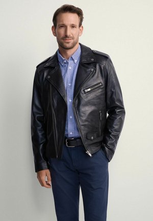 Mężczyzna w czarnej skórzanej kurtce na niebieskiej w kratę koszuli i granatowych spodniach, stojący z jedną ręką w kieszeni na jednolitym tle.