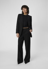Blazer nero su misura e pantaloni a vita alta con top nero corto. Il tessuto appare liscio con linee nette e strutturate. Scarpe nere con punta.