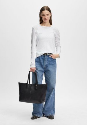Kobieta ubrana w białą koszulę z długim rękawem i szerokie niebieskie dżinsy, trzymająca dużą czarną torbę typu tote, stojąca na jednolitym białym tle.