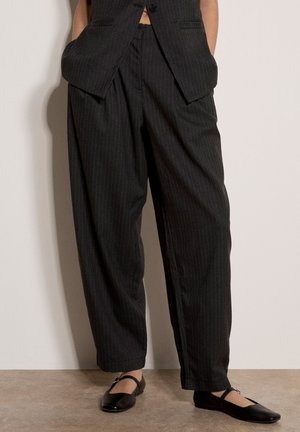 Personne portant un pantalon noir à fines rayures ample et un gilet assorti, les mains dans les poches, associé à des chaussures Mary Jane noires.