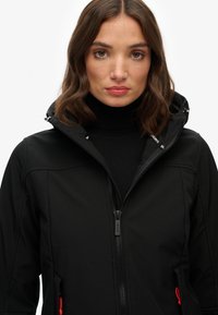 Zwarte softshell jack met capuchon, voorzien van een volledige rits, verstelbare trekkoorden en voortassen met rode accenten.