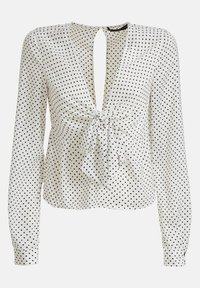 Blouse blanche à pois noirs, manches longues, col en V profond et détail noué à la taille, confectionnée dans un tissu lisse.