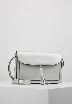 Silberne Umhängetasche mit Klappverschluss und Quasten-Detail, liegend auf einem weißen Podest vor einem schlichten grauen Hintergrund.