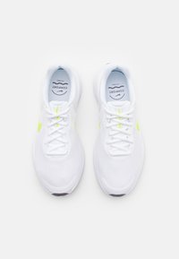 Bílé sportovní boty s síťovaným svrškem, plochými bílými tkaničkami a neonově žlutými akcenty po stranách. Polstrované podrážky pro pohodlí.